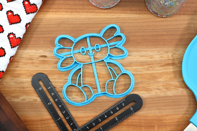 May include: A blue plastic cookie cutter shaped like an axolotl. The axolotl is sitting with its legs crossed and has a happy expression on its face.