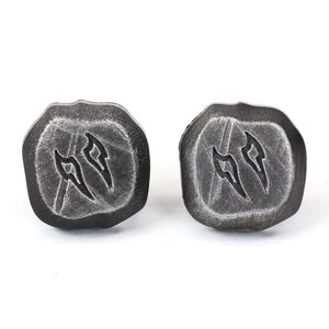 Puede incluir: Un par de pendientes de bot&oacute;n de color gris oscuro, con forma de piedra. Cada pendiente tiene una forma &uacute;nica con un s&iacute;mbolo tallado. Los pendientes est&aacute;n sobre un fondo blanco.