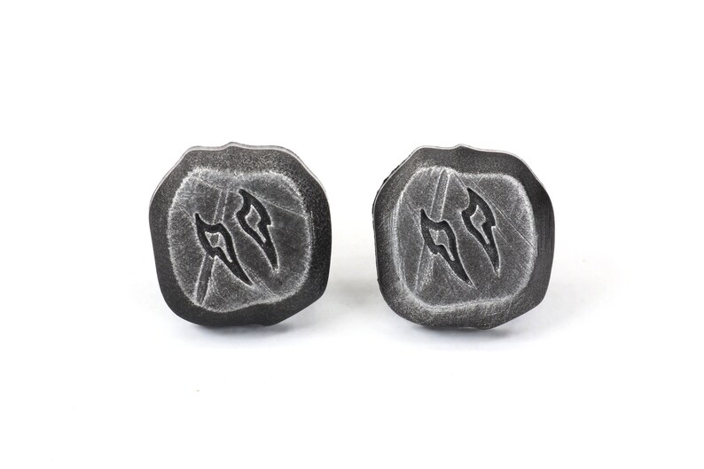 Puede incluir: Un par de pendientes de bot&oacute;n de color gris oscuro, con forma de piedra. Cada pendiente tiene una forma &uacute;nica con un s&iacute;mbolo tallado. Los pendientes est&aacute;n sobre un fondo blanco.