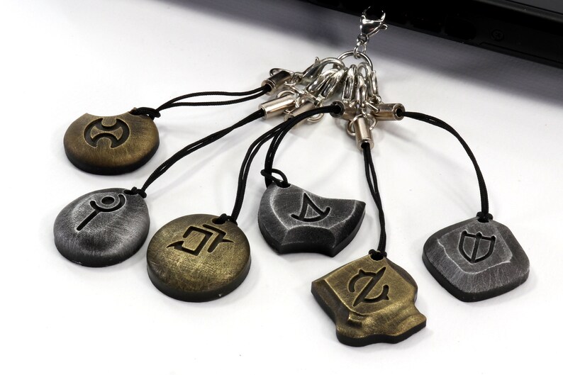 Może przedstawiać: Zestaw siedmiu brelok&oacute;w z r&oacute;żnymi symbolami na każdym. Breloki są wykonane z metalu i mają czarny sznurek. Symbole to koło z linią przechodzącą przez nie, koło z linią przechodzącą przez nie i kropką, koło z linią przechodzącą przez nie i kropką i linią, koło z linią przechodzącą przez nie i kropką i linią i k&oacute;łkiem, koło z linią przechodzącą przez nie i kropką i linią i k&oacute;łkiem i linią, koło z linią przechodzącą przez nie i kropką i linią i k&oacute;łkiem i linią i k&oacute;łkiem, oraz koło z linią przechodzącą przez nie i kropką i linią i k&oacute;łkiem i linią i k&oacute;łkiem i linią.