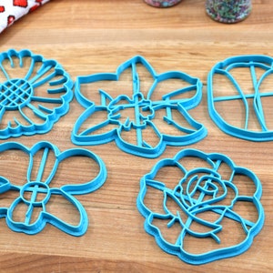May include: Five blue plastic cookie cutters shaped like different flowers. The cutters are on a wooden surface.