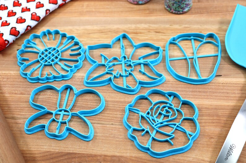 May include: Five blue plastic cookie cutters shaped like different flowers. The cutters are on a wooden surface.