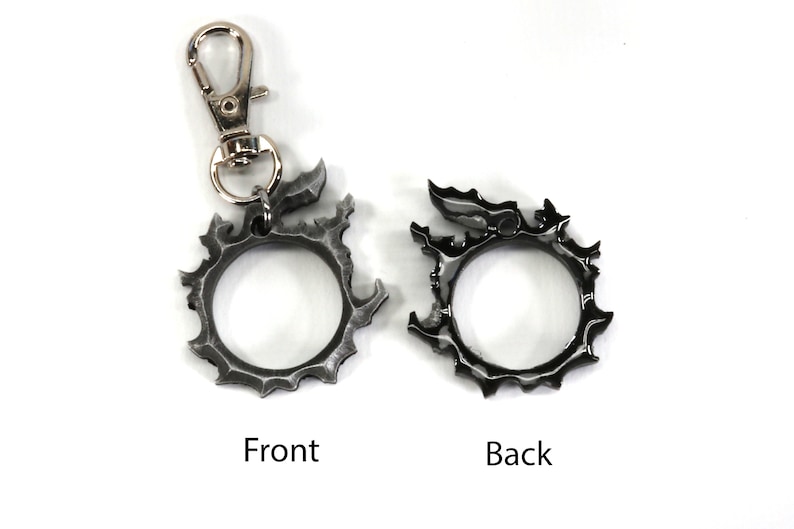 Puede incluir: Un llavero de metal plateado con un dise&ntilde;o circular que presenta una cabeza de drag&oacute;n estilizada. El llavero tiene un cierre de metal plateado y se muestra desde el frente y la parte posterior.