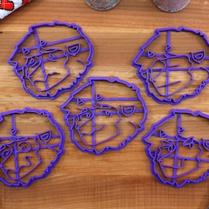 May include: Five purple plastic cookie cutters shaped like a cartoon dog's face. The cutters are on a wooden surface.
