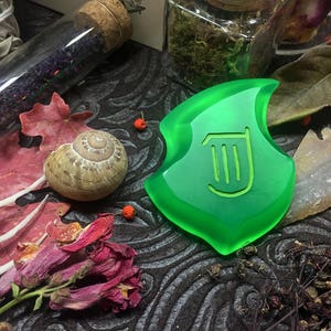 May include: A green glass pendant with a harp symbol in the center. The pendant is surrounded by various objects, including a snail shell, dried flowers, and a glass vial.