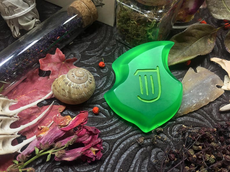 May include: A green glass pendant with a harp symbol in the center. The pendant is surrounded by various objects, including a snail shell, dried flowers, and a glass vial.