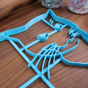 May include: A blue plastic cookie cutter shaped like a cat's face. The cutter has detailed features, including eyes, a nose, and whiskers.