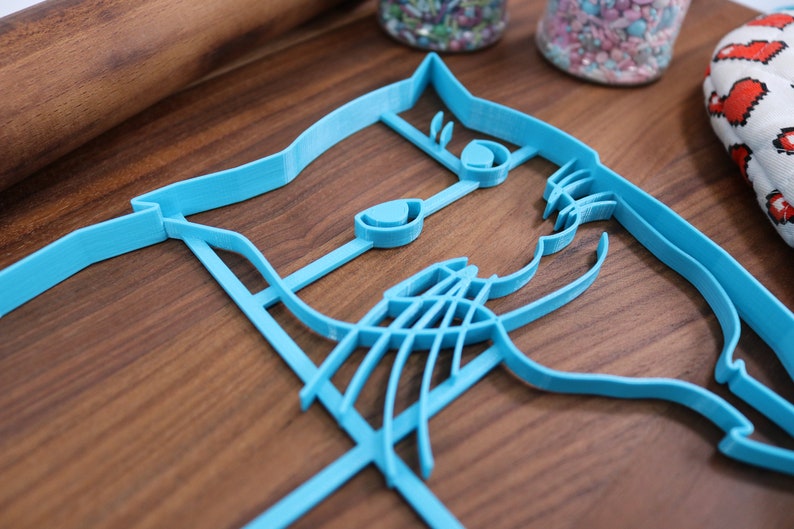 May include: A blue plastic cookie cutter shaped like a cat's face. The cutter has detailed features, including eyes, a nose, and whiskers.