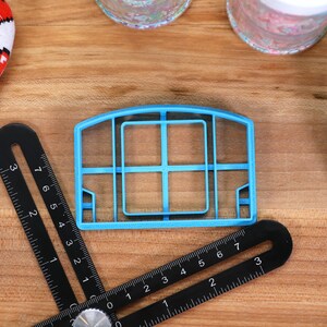 May include: A blue plastic cookie cutter shaped like a basketball hoop with a net. The cutter is on a wooden surface with a black ruler.