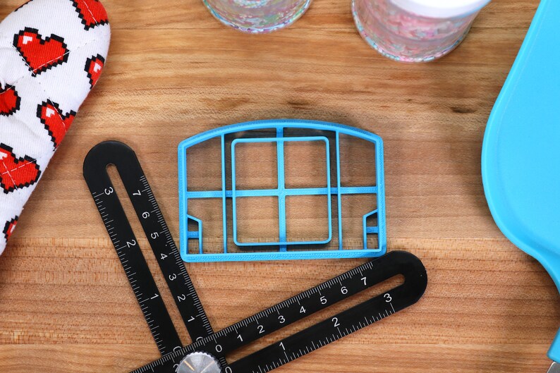 May include: A blue plastic cookie cutter shaped like a basketball hoop with a net. The cutter is on a wooden surface with a black ruler.