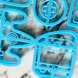 May include: A set of five blue plastic cookie cutters shaped like video game controllers and a game cartridge.