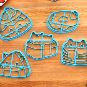 May include: Five blue plastic cookie cutters shaped like different types of sushi with cute cat faces. The cutters are on a wooden surface.
