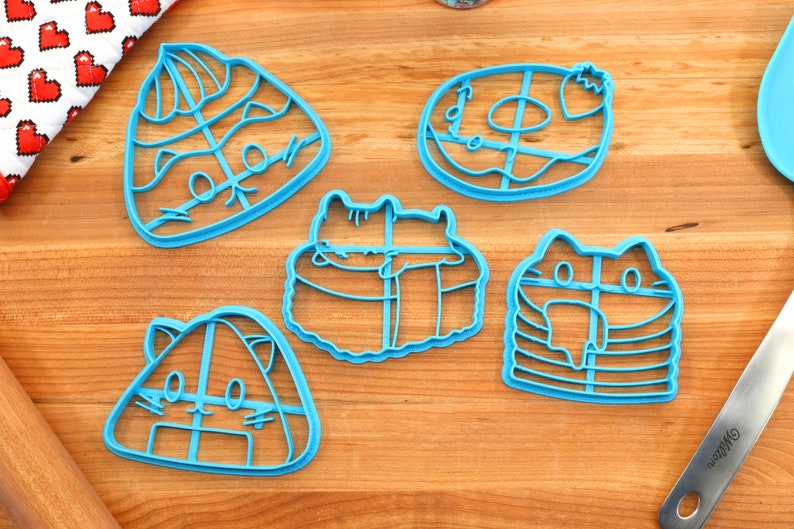 May include: Five blue plastic cookie cutters shaped like different types of sushi with cute cat faces. The cutters are on a wooden surface.