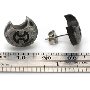 May include: A pair of silver stud earrings with a crescent moon design and a symbol in the centre. The earrings are shown next to a ruler, measuring approximately 1 cm in diameter.