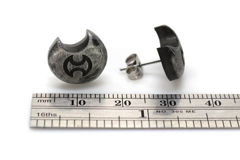 May include: A pair of silver stud earrings with a crescent moon design and a symbol in the centre. The earrings are shown next to a ruler, measuring approximately 1 cm in diameter.
