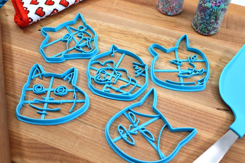 May include: Five blue plastic cookie cutters shaped like different animal faces. The cutters are on a wooden surface.