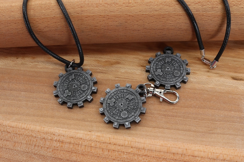 May include: Three silver-colored pendants with a detailed design, two on black cords and one on a silver chain. The pendants are shaped like gears with a circular design in the center.