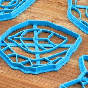 May include: Four blue plastic cookie cutters shaped like leaves. The cutters have intricate designs and are arranged on a wooden surface.