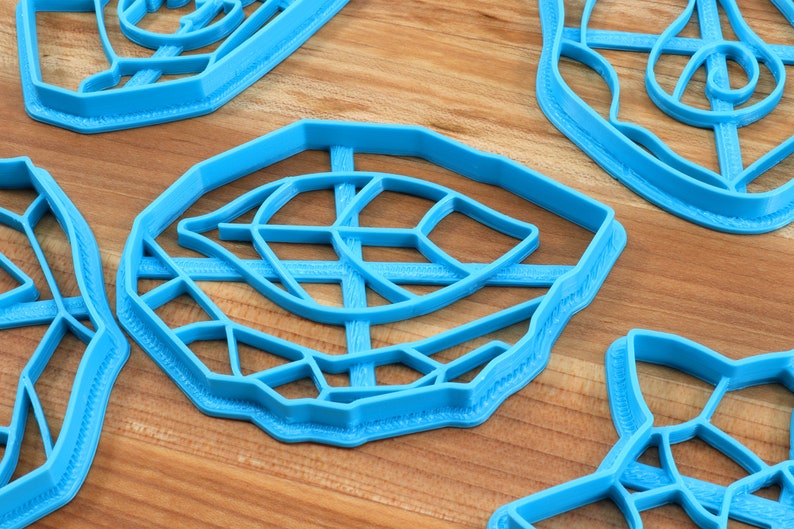 May include: Four blue plastic cookie cutters shaped like leaves. The cutters have intricate designs and are arranged on a wooden surface.