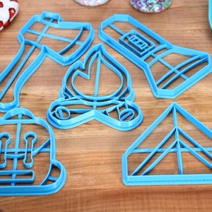 Op de afbeelding: Een set van vijf blauwe plastic koekjesvormen in de vorm van een zaklamp, een bijl, een kampvuur, een rugzak en een tent.