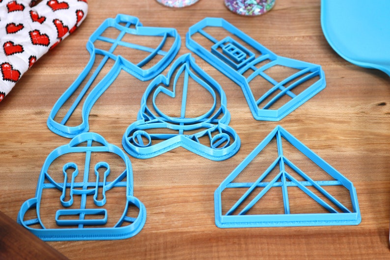 Op de afbeelding: Een set van vijf blauwe plastic koekjesvormen in de vorm van een zaklamp, een bijl, een kampvuur, een rugzak en een tent.