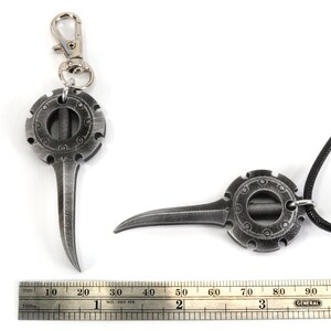 May include: A silver-colored metal pendant with a circular design and a pointed tip, attached to a black cord. The pendant is also shown with a silver-colored metal key chain.