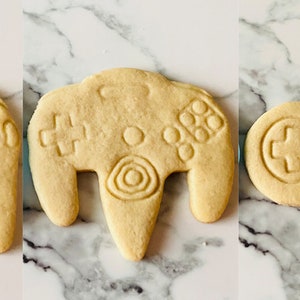 May include: Five different cookie cutters shaped like video game controllers. The cutters are made of a light brown dough and are ready to be baked.