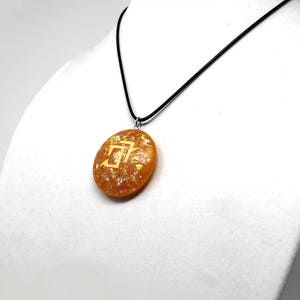 May include: A gold pendant necklace with a round, orange resin pendant featuring a gold symbol. The pendant is on a black cord.