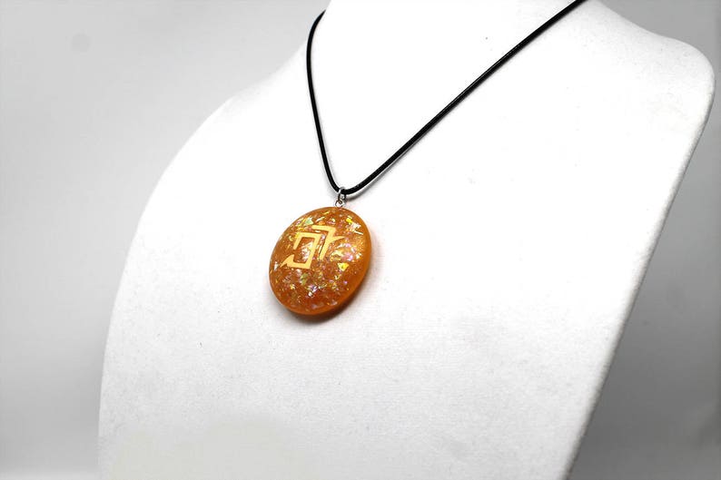 May include: A gold pendant necklace with a round, orange resin pendant featuring a gold symbol. The pendant is on a black cord.