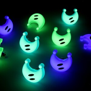 May include: Nine glow-in-the-dark plastic moon-shaped buttons in various shades of green, blue, and purple. The buttons have two black dots on each.