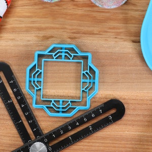 May include: A blue plastic cookie cutter shaped like a square with an intricate design. The cutter is on a wooden surface with a ruler and a blue spatula.
