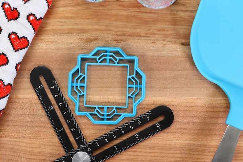 May include: A blue plastic cookie cutter shaped like a square with an intricate design. The cutter is on a wooden surface with a ruler and a blue spatula.