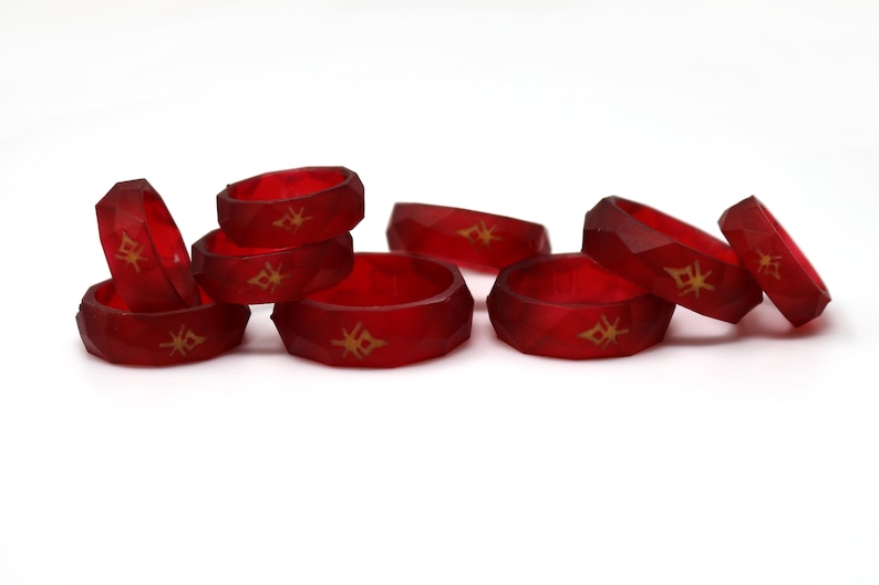 May include: A group of eight red silicone rings with a gold geometric design. The rings are stacked and scattered on a white surface.