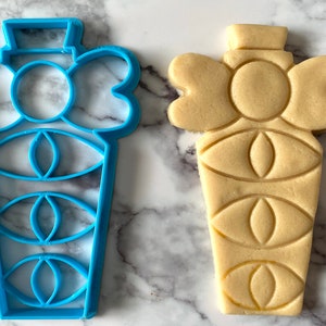 Pode incluir: Um cortador de biscoitos de pl&aacute;stico azul em forma de frasco de perfume com um la&ccedil;o no topo. Ao lado dele est&aacute; um biscoito de a&ccedil;&uacute;car marrom claro cortado com a mesma forma.