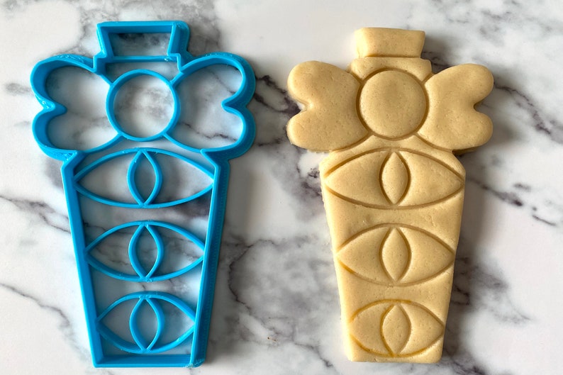 Pode incluir: Um cortador de biscoitos de pl&aacute;stico azul em forma de frasco de perfume com um la&ccedil;o no topo. Ao lado dele est&aacute; um biscoito de a&ccedil;&uacute;car marrom claro cortado com a mesma forma.