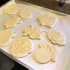May include: A baking sheet with parchment paper and nine unbaked sugar cookies. The cookies are shaped like various characters, including a mushroom, a star, and a turtle.