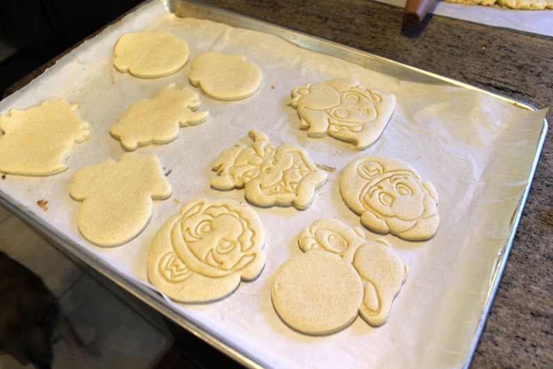 May include: A baking sheet with parchment paper and nine unbaked sugar cookies. The cookies are shaped like various characters, including a mushroom, a star, and a turtle.