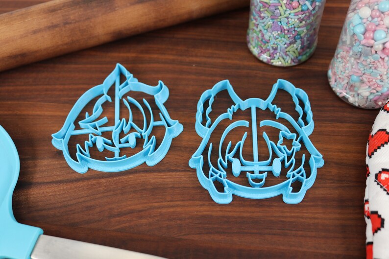 May include: Two blue plastic cookie cutters shaped like cartoon characters. The cutters are on a wooden surface.