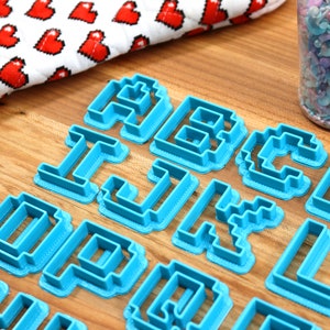 May include: A set of blue plastic cookie cutters shaped like letters of the alphabet in a pixelated style. The cutters are arranged on a wooden surface.