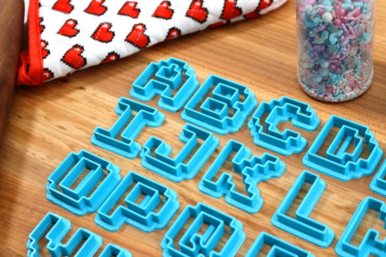 May include: A set of blue plastic cookie cutters shaped like letters of the alphabet in a pixelated style. The cutters are arranged on a wooden surface.