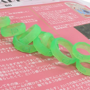 May include: A close-up of a pile of bright green silicone rings with the letter "B" embossed on each ring. The rings are on a pink and white surface with Japanese text.
