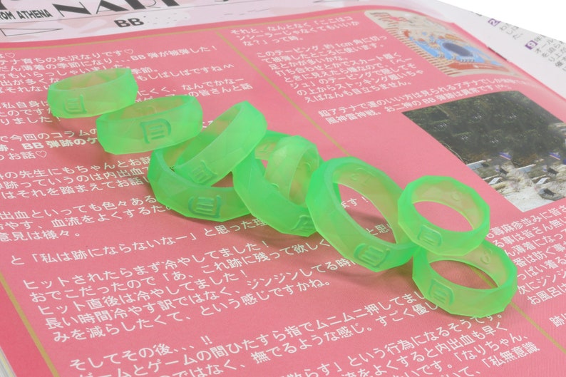 May include: A close-up of a pile of bright green silicone rings with the letter "B" embossed on each ring. The rings are on a pink and white surface with Japanese text.