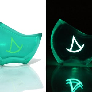 May include: A teal blue resin paperweight with a green glow-in-the-dark symbol of a sailboat inside. The paperweight is shaped like a crescent moon.