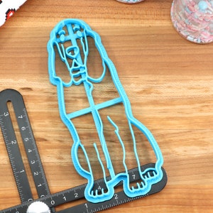 May include: A light blue plastic cookie cutter shaped like a standing dog. The cutter is on a wooden surface with a ruler and a jar of sprinkles in the background.
