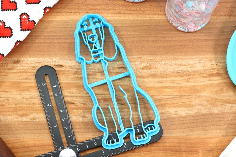 May include: A light blue plastic cookie cutter shaped like a standing dog. The cutter is on a wooden surface with a ruler and a jar of sprinkles in the background.