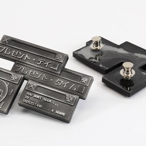 Op de afbeelding: Een set van drie zilverkleurige metalen pinnen met een cartoonfiguur en tekst in het Japans die "Cadeau - Tijd" luidt. De pinnen zijn bevestigd aan zwarte achtergronden met zilverkleurige metalen pinruggen.