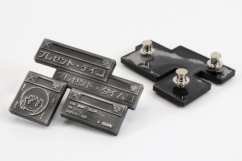 Op de afbeelding: Een set van drie zilverkleurige metalen pinnen met een cartoonfiguur en tekst in het Japans die "Cadeau - Tijd" luidt. De pinnen zijn bevestigd aan zwarte achtergronden met zilverkleurige metalen pinruggen.