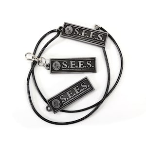 May include: Three black metal rectangular tags with the text "S.E.E.S." and a small logo on each tag. The tags are attached to a black cord.