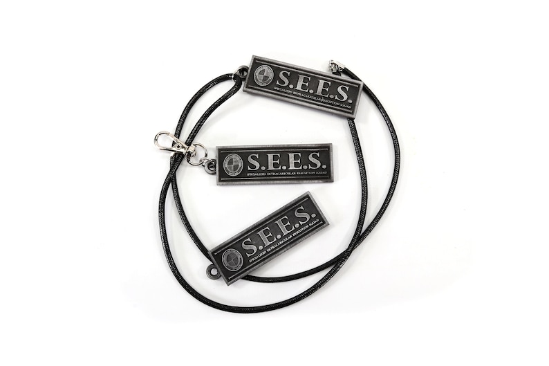May include: Three black metal rectangular tags with the text "S.E.E.S." and a small logo on each tag. The tags are attached to a black cord.