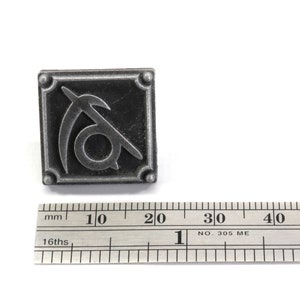 May include: A silver metal pin with a stylized symbol of a sickle and a circle. The pin is about 1.5 inches wide and is shown next to a ruler for scale.